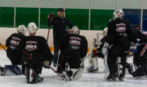 Coach giving instructions to ice hockey players in goalie gear during practice.
